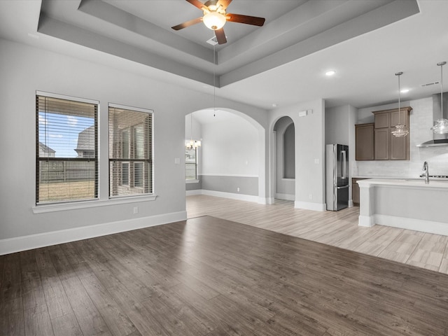 unfurnished living room with a raised ceiling, light wood-style flooring, ceiling fan with notable chandelier, arched walkways, and baseboards