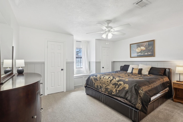 bedroom with a ceiling fan, a wainscoted wall, visible vents, a textured ceiling, and light colored carpet