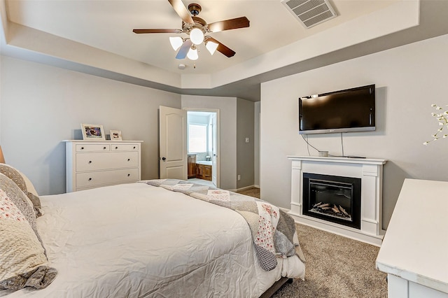 carpeted bedroom featuring visible vents, a ceiling fan, ensuite bathroom, a glass covered fireplace, and a raised ceiling