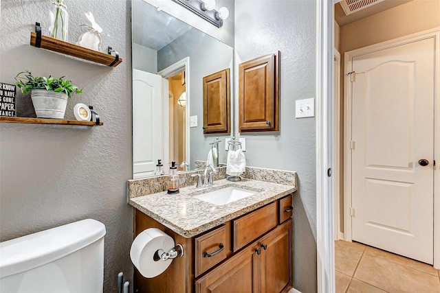 half bathroom with tile patterned floors, visible vents, toilet, vanity, and a textured wall