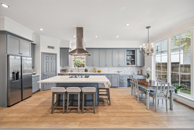 kitchen featuring gray cabinets, light countertops, stainless steel refrigerator with ice dispenser, tasteful backsplash, and island range hood