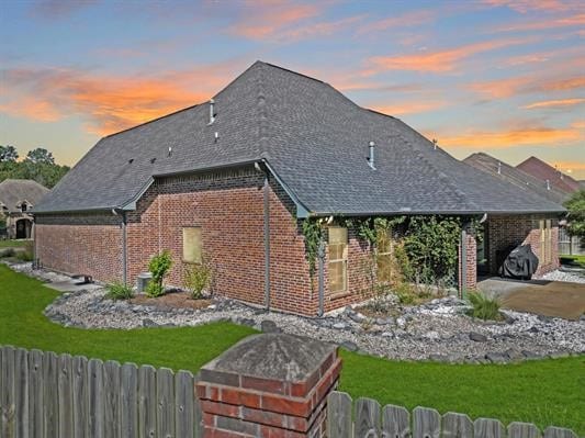 back of property at dusk featuring a yard, brick siding, a shingled roof, and fence