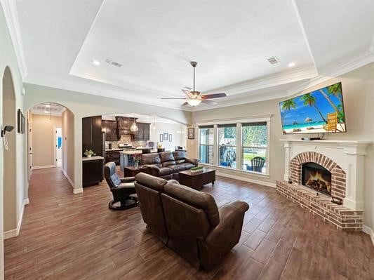 living room featuring dark wood finished floors, a fireplace, arched walkways, ornamental molding, and a raised ceiling