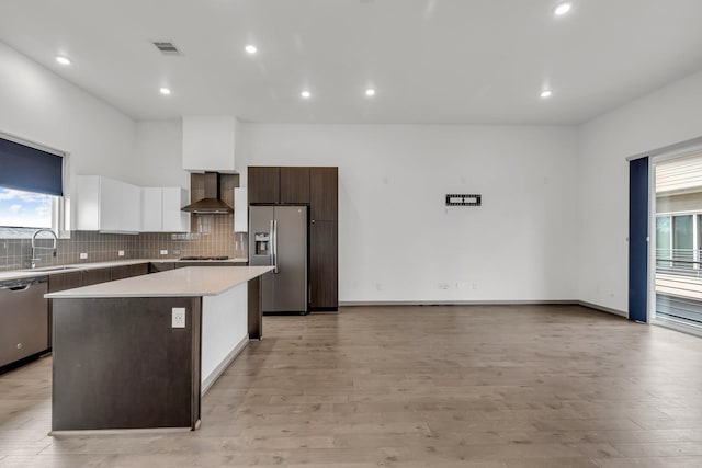 kitchen featuring a kitchen island, a sink, appliances with stainless steel finishes, wall chimney exhaust hood, and tasteful backsplash