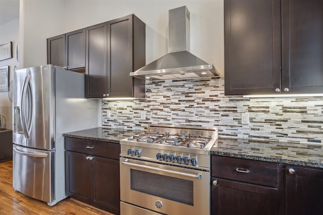 kitchen featuring dark stone counters, appliances with stainless steel finishes, wall chimney range hood, decorative backsplash, and dark brown cabinets