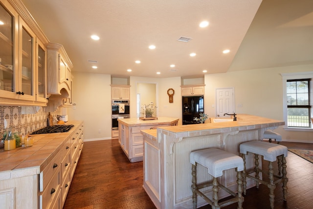 kitchen featuring tile counters, dark wood-style floors, a kitchen island with sink, and black appliances