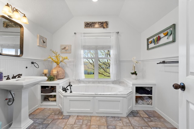 full bathroom with vaulted ceiling, stone finish flooring, a bath, and wainscoting