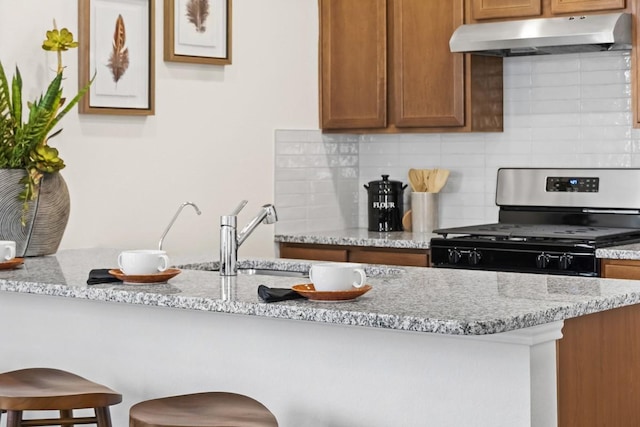 kitchen with under cabinet range hood, decorative backsplash, stainless steel gas stove, and light stone counters