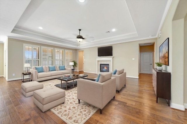 living area featuring a glass covered fireplace, ceiling fan, light wood-type flooring, and a tray ceiling