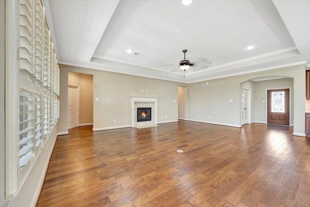 unfurnished living room with a tray ceiling, arched walkways, dark wood finished floors, and a ceiling fan