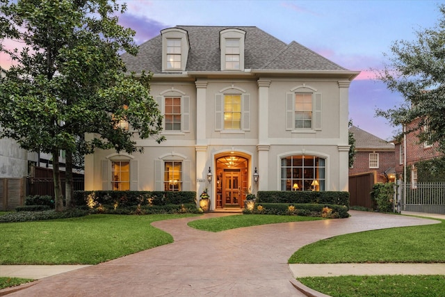 french country home featuring fence, roof with shingles, stucco siding, a lawn, and decorative driveway