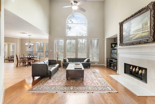 living area featuring baseboards, ornamental molding, a tiled fireplace, ceiling fan with notable chandelier, and light wood-type flooring