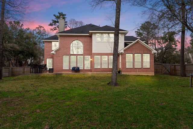 back of house at dusk featuring a lawn, brick siding, a fenced backyard, and a chimney