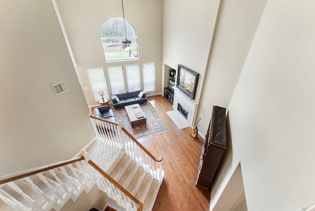 living room featuring a ceiling fan, visible vents, light wood finished floors, a high ceiling, and a fireplace with flush hearth