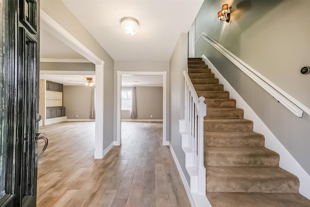 foyer entrance featuring a ceiling fan, baseboards, light wood finished floors, ornamental molding, and stairs