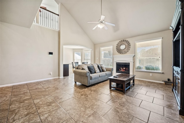 living area featuring baseboards, a lit fireplace, arched walkways, high vaulted ceiling, and a ceiling fan