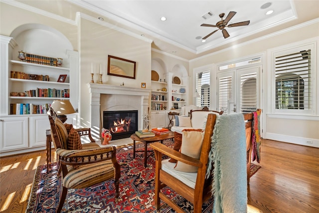 living area featuring visible vents, built in shelves, light wood-style flooring, a glass covered fireplace, and a raised ceiling