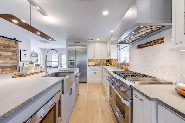 kitchen featuring premium appliances, tasteful backsplash, white cabinetry, a barn door, and wall chimney range hood