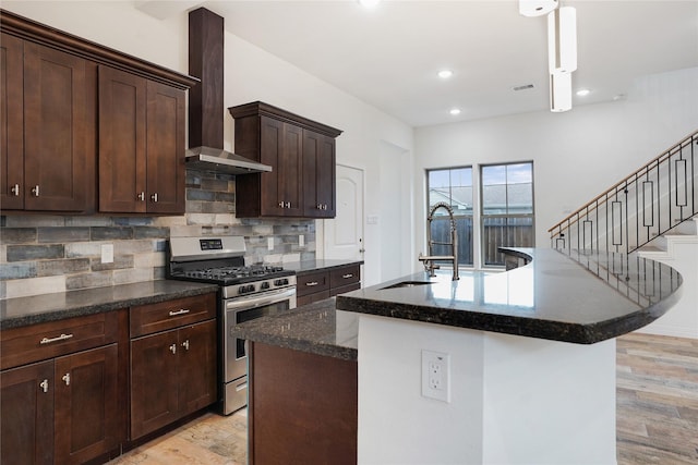 kitchen with stainless steel range with gas cooktop, an island with sink, a sink, wall chimney exhaust hood, and light wood-type flooring