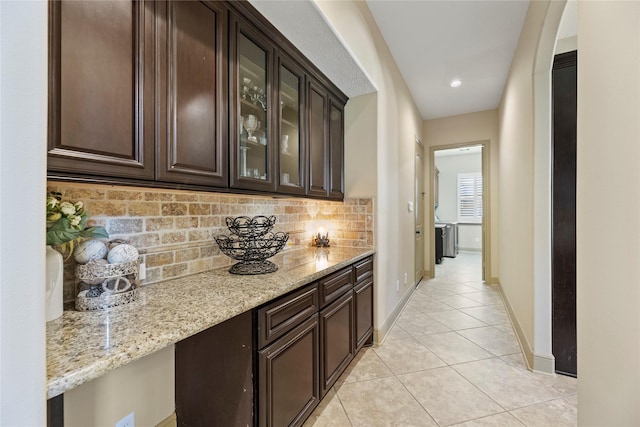 interior space featuring light stone counters, glass insert cabinets, light tile patterned flooring, decorative backsplash, and dark brown cabinets