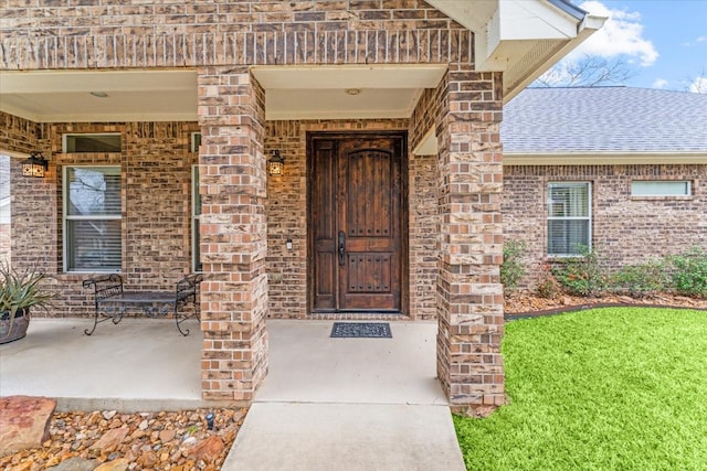 property entrance featuring a yard, brick siding, a porch, and a shingled roof