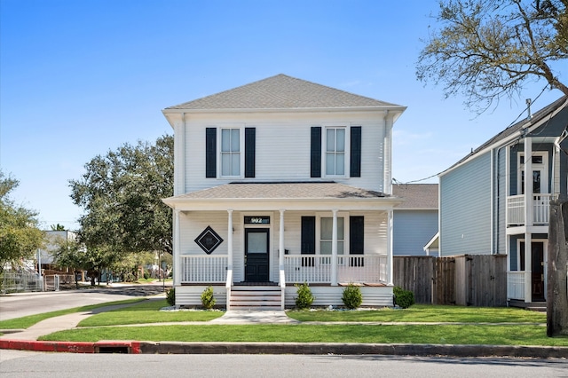 american foursquare style home with covered porch, a shingled roof, a front yard, and fence