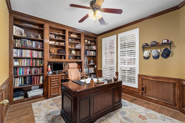 home office with light wood-type flooring, a wainscoted wall, a ceiling fan, a textured ceiling, and crown molding