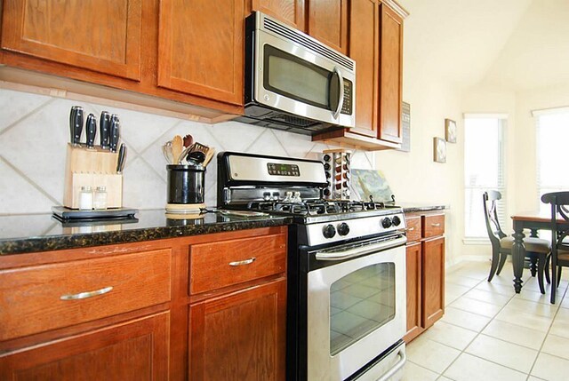 kitchen with brown cabinets, dark stone countertops, backsplash, stainless steel appliances, and light tile patterned floors