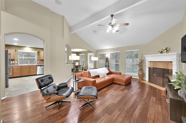 living room with light wood-type flooring, beam ceiling, high vaulted ceiling, a ceiling fan, and a tile fireplace