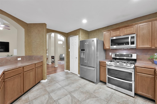 kitchen featuring brown cabinetry, arched walkways, stainless steel appliances, light countertops, and tasteful backsplash