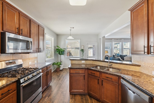 kitchen with a sink, dark stone counters, appliances with stainless steel finishes, brown cabinetry, and dark wood-style flooring