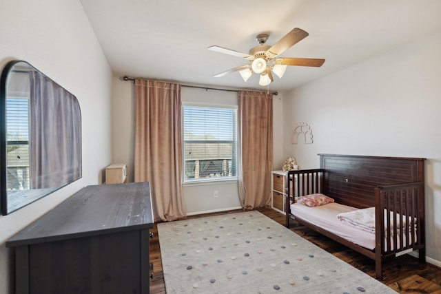 bedroom featuring dark wood finished floors, a nursery area, baseboards, and ceiling fan