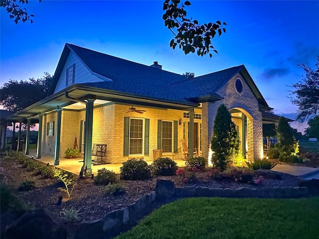 property exterior at dusk featuring brick siding, ceiling fan, covered porch, a lawn, and a chimney