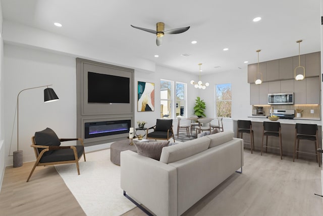 living area featuring visible vents, light wood-style flooring, recessed lighting, a glass covered fireplace, and ceiling fan with notable chandelier