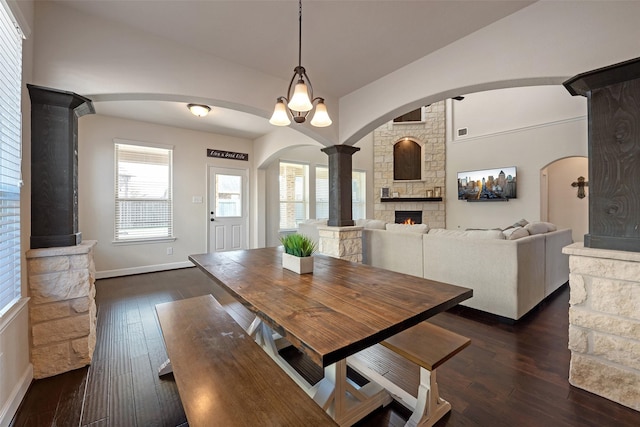 dining area featuring baseboards, dark wood finished floors, decorative columns, a stone fireplace, and vaulted ceiling