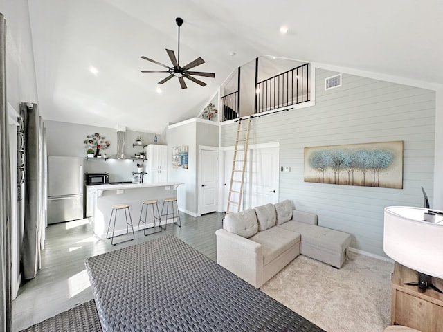 living area with visible vents, high vaulted ceiling, a ceiling fan, wooden walls, and baseboards