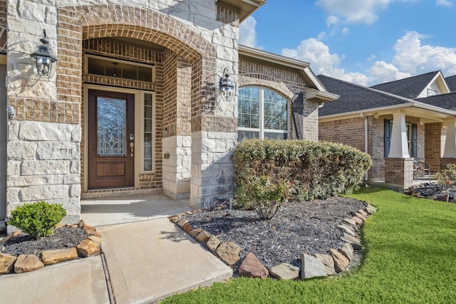 doorway to property with stone siding, brick siding, a lawn, and a shingled roof