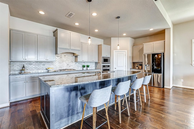 kitchen with light stone counters, a center island with sink, visible vents, dark wood-style flooring, and stainless steel appliances