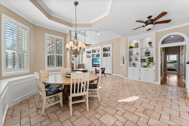 dining area featuring a wainscoted wall, ceiling fan with notable chandelier, built in features, crown molding, and a raised ceiling