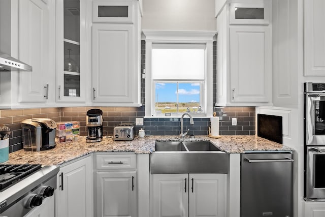kitchen featuring backsplash, glass insert cabinets, wall chimney range hood, white cabinetry, and a sink