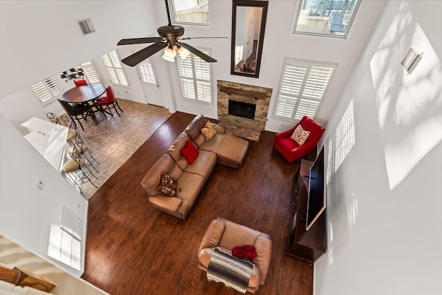 living area with wood finished floors, visible vents, a ceiling fan, a high ceiling, and a stone fireplace