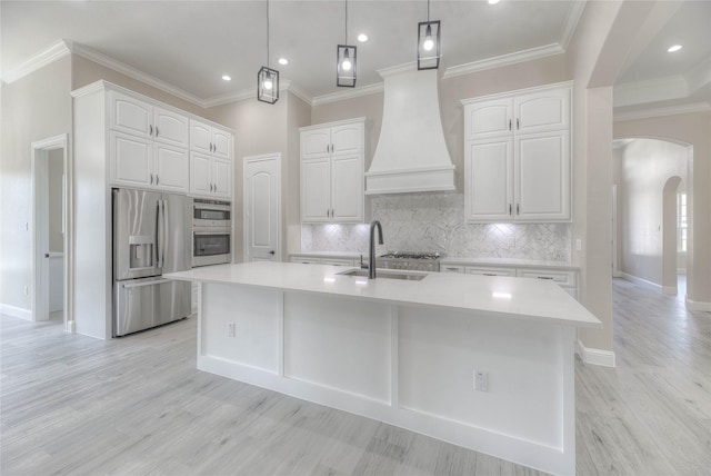 kitchen featuring an island with sink, arched walkways, a sink, stainless steel appliances, and white cabinetry