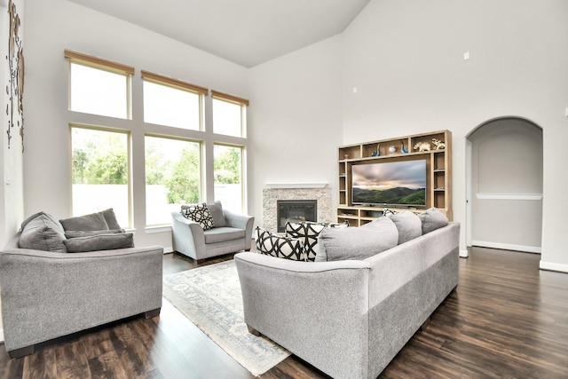 living area with arched walkways, a high ceiling, a stone fireplace, baseboards, and dark wood-style flooring