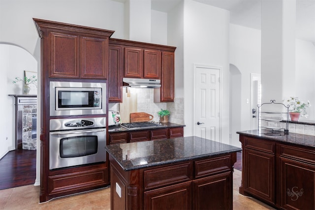 kitchen with under cabinet range hood, backsplash, stainless steel appliances, arched walkways, and dark stone counters