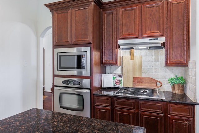 kitchen with dark stone counters, arched walkways, decorative backsplash, under cabinet range hood, and appliances with stainless steel finishes