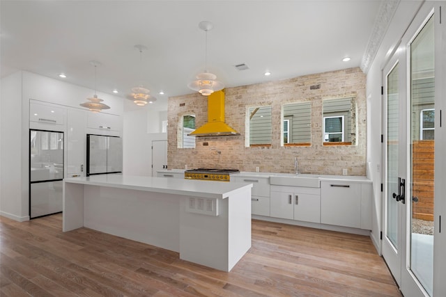 kitchen with light wood-style flooring, stainless steel refrigerator, stove, white cabinetry, and wall chimney range hood