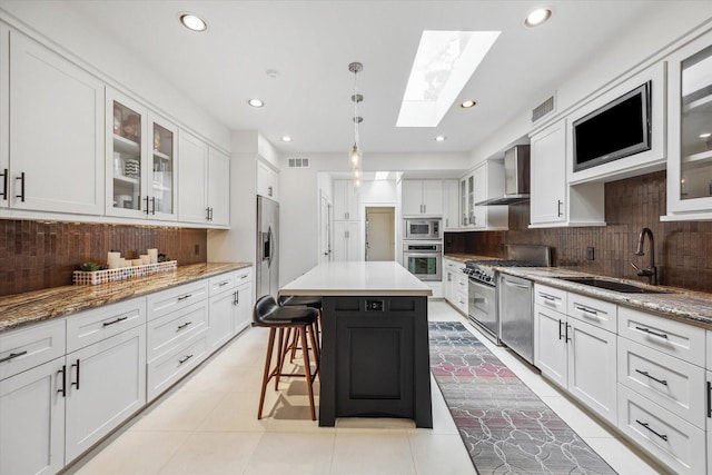 kitchen with wall chimney range hood, a skylight, white cabinets, stainless steel appliances, and a sink