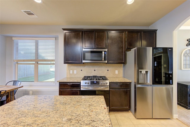 kitchen with light stone countertops, arched walkways, stainless steel appliances, dark brown cabinetry, and backsplash