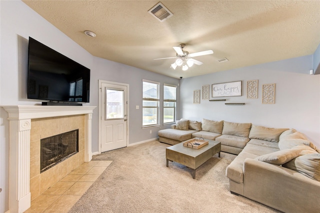 living room with a ceiling fan, visible vents, a textured ceiling, a tiled fireplace, and light carpet