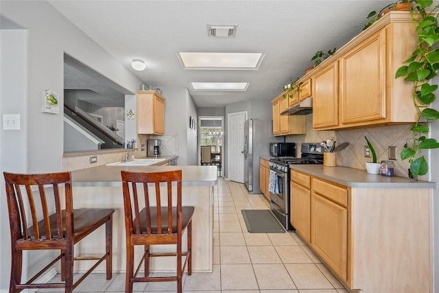 kitchen with under cabinet range hood, light brown cabinetry, a breakfast bar area, light tile patterned flooring, and stainless steel appliances